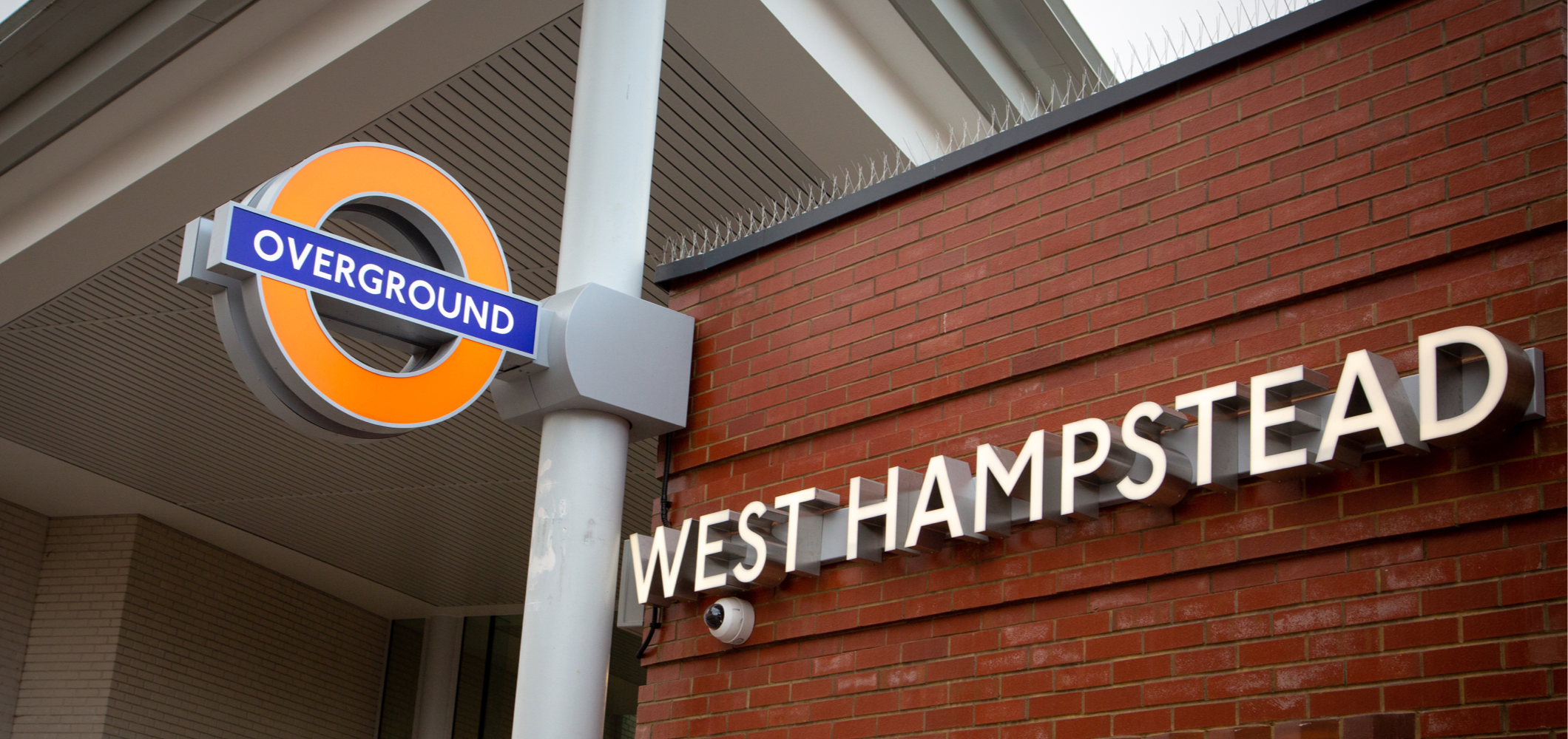A photo of signage at the entrance of West Hampstead Overground station: a 3D hanging sign of the London Overground logo, next to a brick wall displaying the name of the station in 3D metal typography.
