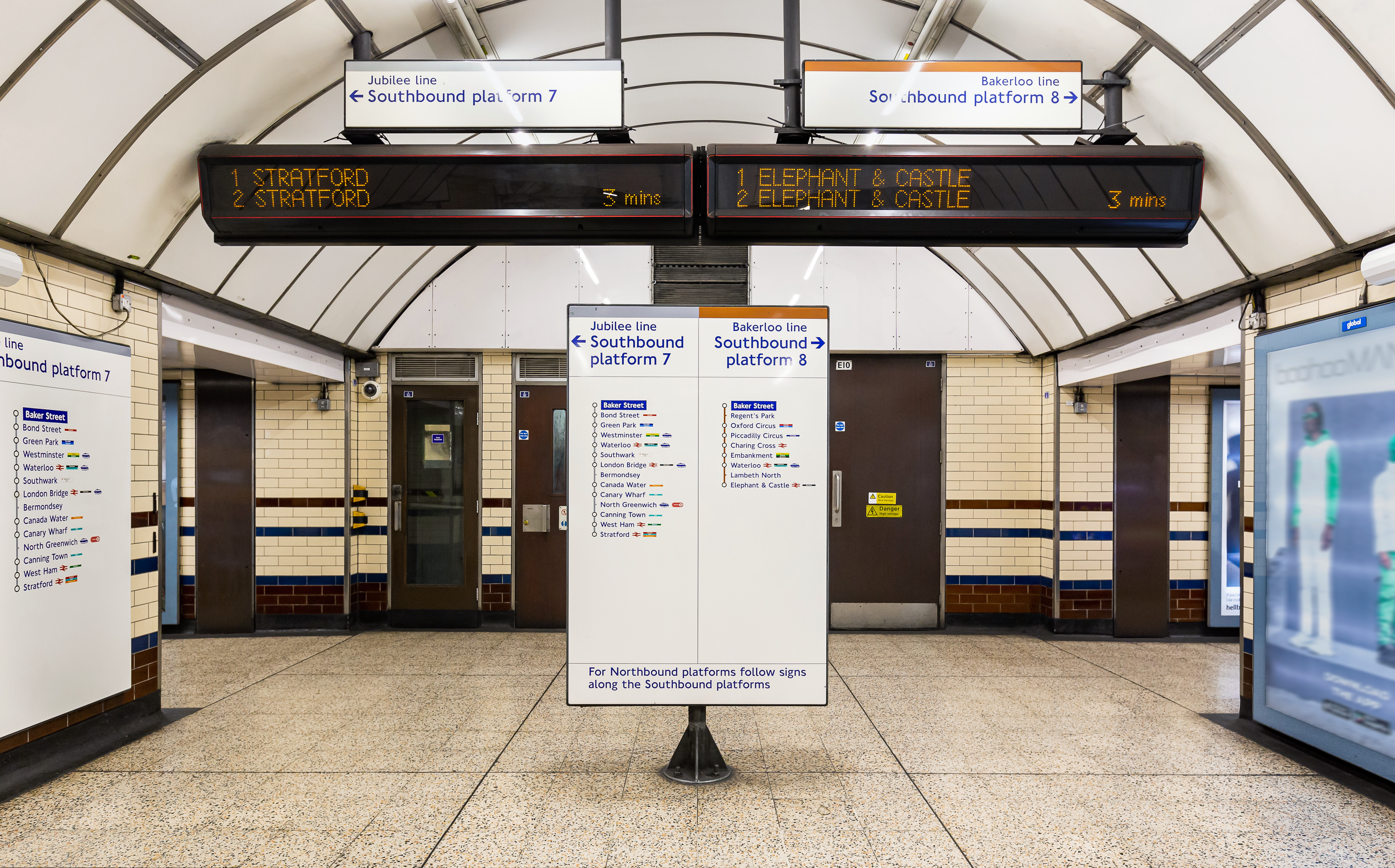 A photography of a concourse in an underground tube station. There are signs indicating two lines in two opposing directions, and screens showing live departures for each line.