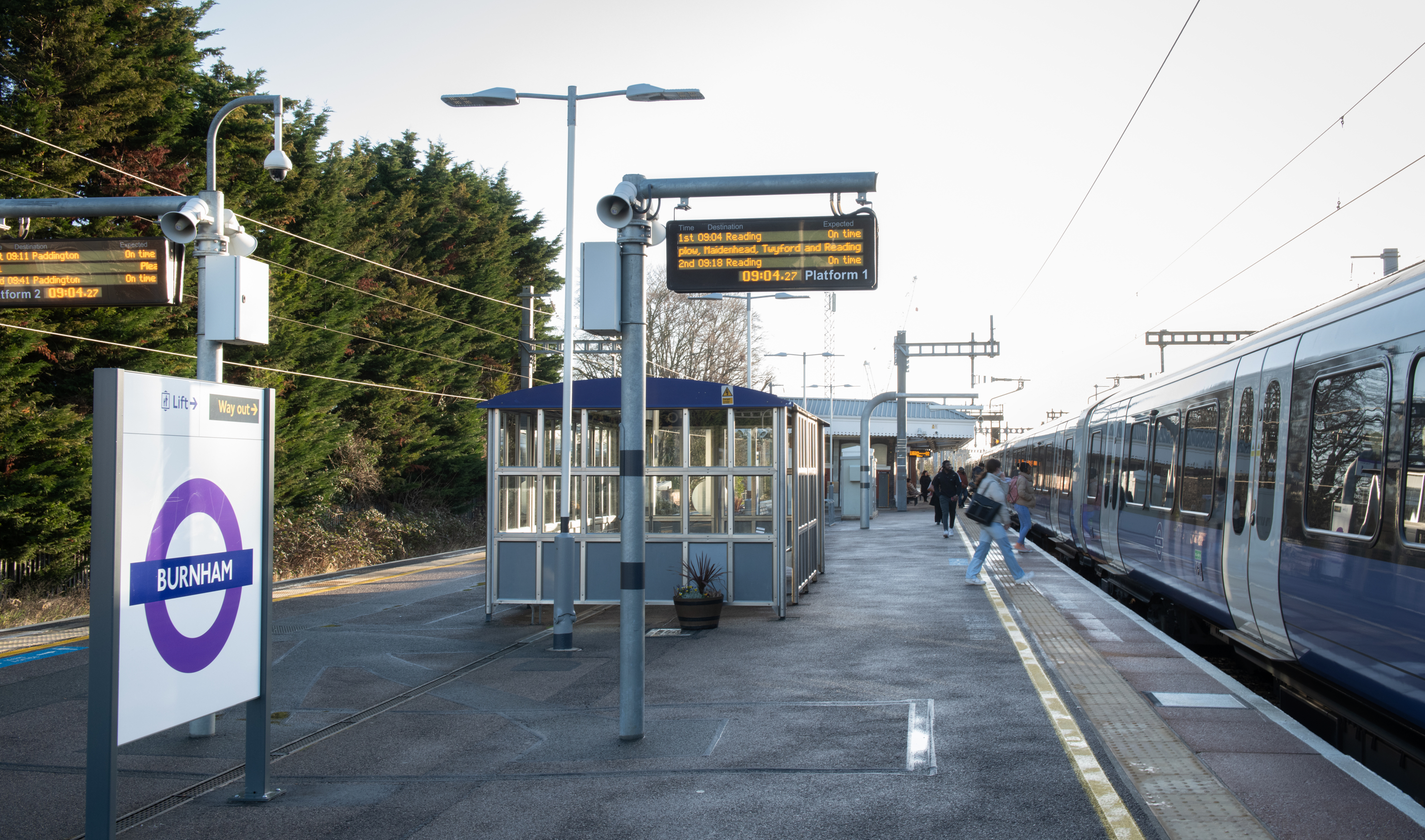 Photograph of an Elizabeth line outdoor platform. An led screen is displaying departure information. A train is at the platform and a few passengers are preparing to board.