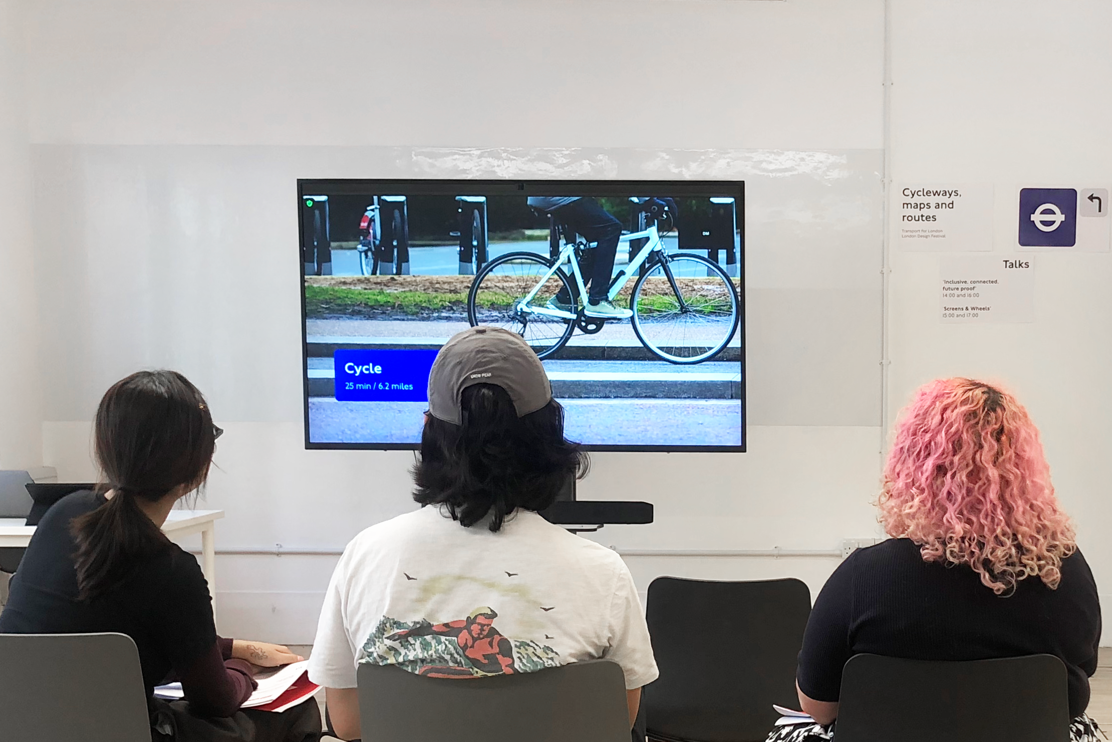 A photograph of three seated visiors watching a presentation about cycling on a large screen